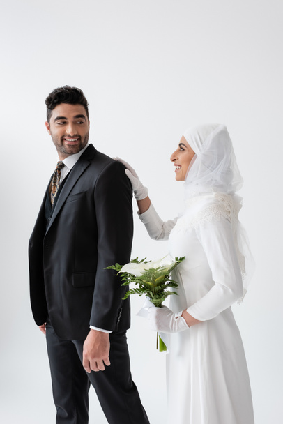 happy muslim bride in wedding dress holding bouquet of calla lily and standing behind groom in suit isolated on grey - Foto, Imagem
