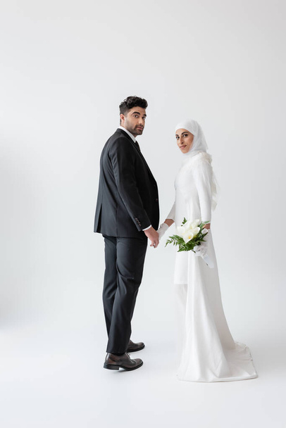 usměvavá muslimská nevěsta v bílých šatech s kyticí leknínů držících se za ruce s ženichem na šedi - Fotografie, Obrázek