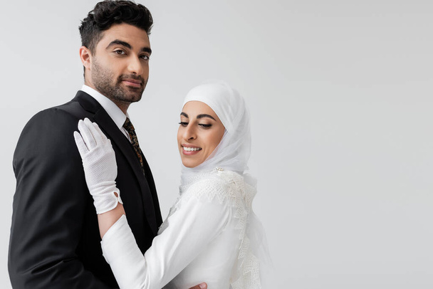 happy muslim bride in hijab and wedding dress and groom hugging isolated on grey - Fotoğraf, Görsel