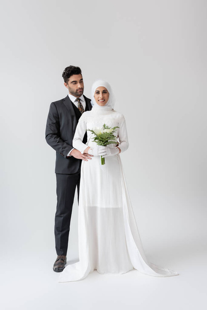 muslimský ženich objímající šťastnou nevěstu ve svatebních šatech s kyticí na šedé  - Fotografie, Obrázek