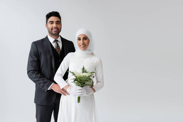 Мусульманский жених обнимает веселую невесту в свадебном платье с букетом, изолированным на сером  - Фото, изображение