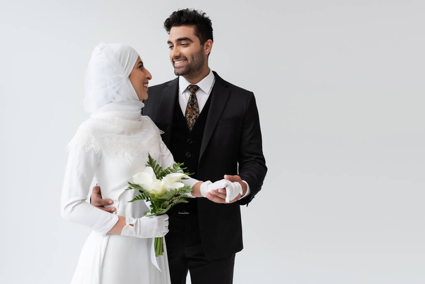 muslim groom holding hand of smiling bride in wedding dress with bouquet isolated on grey  - Foto, imagen