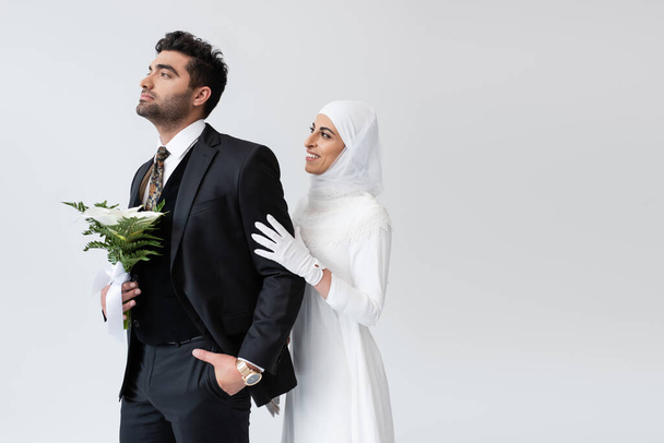 happy muslim bride in hijab hugging groom with bouquet isolated on grey  - Foto, Imagem