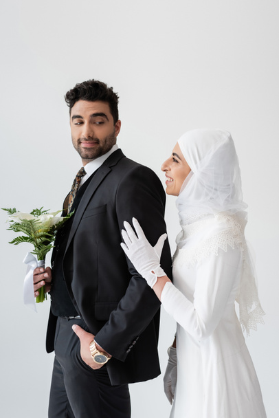 happy muslim bride in hijab hugging groom with bouquet of calla lily flowers isolated on grey  - Foto, Imagem