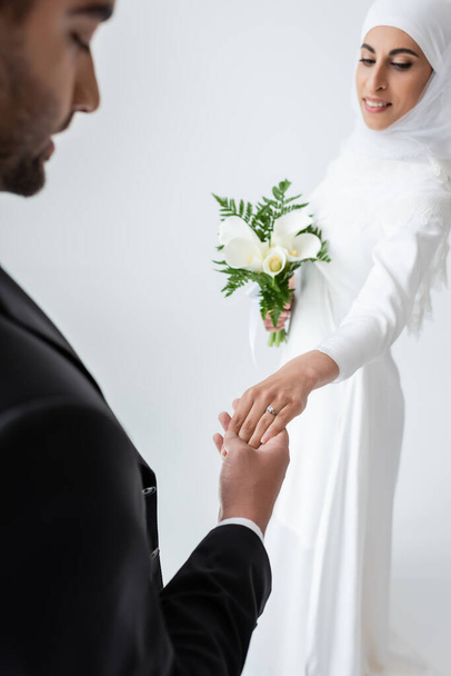 happy muslim bride with bouquet holding hands with blurred groom isolated on grey  - Photo, Image