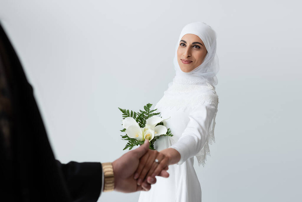 glückliche muslimische Braut mit Strauß Händchen haltend mit Bräutigam isoliert auf grau  - Foto, Bild