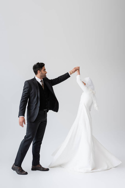 full length of muslim groom in suit and bride in white dress dancing on grey  - Zdjęcie, obraz