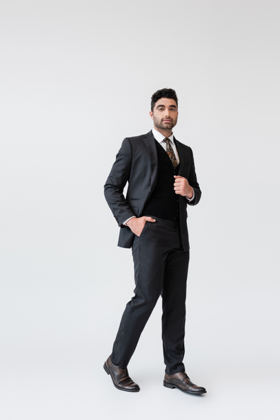 full length of muslim groom in formal wear standing with hand in pocket on grey  - 写真・画像