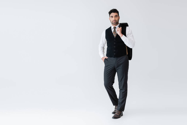 full length of muslim man in vest standing with hand in pocket and holding blazer isolated on grey - Fotoğraf, Görsel