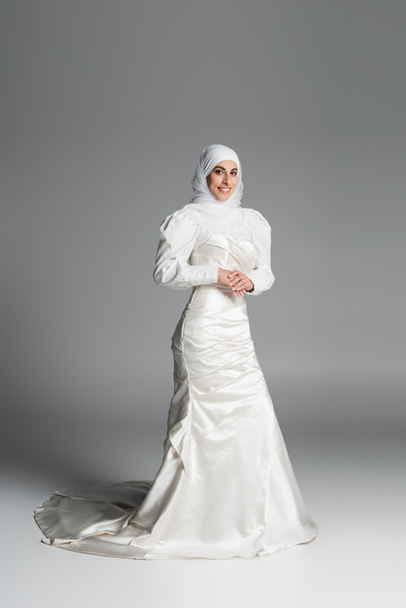full length of happy muslim bride in wedding dress and hijab standing on dark grey  - Фото, зображення