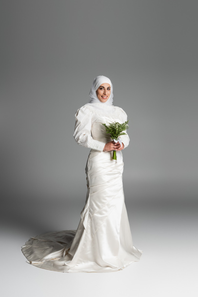 full length of pleased muslim bride in white dress and hijab holding wedding bouquet on dark grey  - Foto, imagen