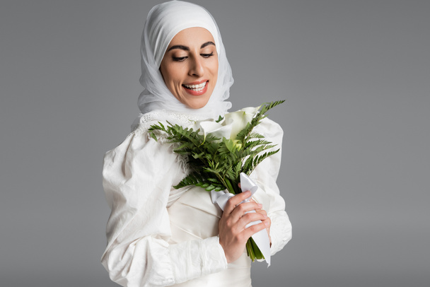 happy muslim bride in white dress and hijab looking at wedding bouquet on dark grey  - Foto, immagini