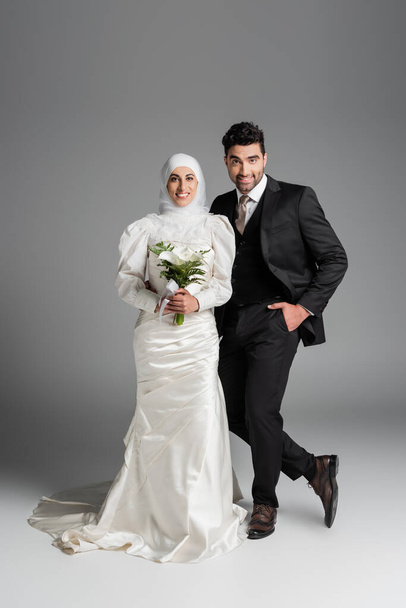счастливый жених в костюме позирует рядом с мусульманской невестой со свадебным букетом на сером - Фото, изображение