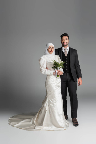 full length of happy groom in suit near muslim bride with wedding bouquet on grey - Photo, Image
