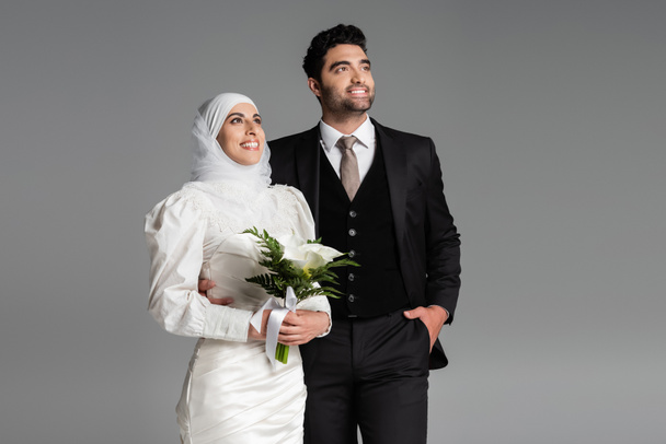 Bräutigam im Anzug posiert neben muslimischer glücklicher Braut mit Brautstrauß auf grau - Foto, Bild