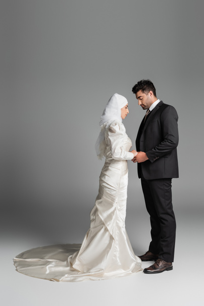 full length of muslim bride in wedding dress and groom in suit holding hands on grey - Foto, Imagem