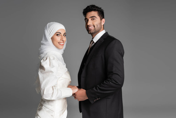 happy muslim bride in wedding dress and groom in suit holding hands isolated on grey - 写真・画像