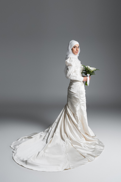 full length view of muslim bride in gorgeous wedding dress standing with bouquet on grey - Фото, изображение