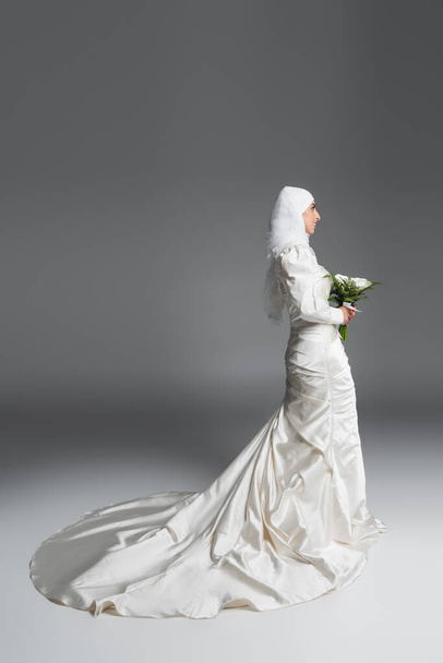 side view of happy muslim woman in gorgeous wedding dress standing with bouquet on grey - Photo, image