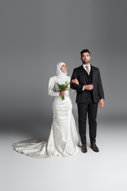 longueur totale du marié en costume debout avec mariée musulmane heureuse avec bouquet de mariage de fleurs de lys calla sur gris - Photo, image