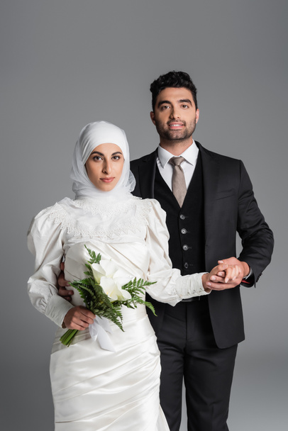 marié heureux en costume tenant la main de mariée musulmane avec bouquet de mariage de fleurs de lys calla isolé sur gris - Photo, image