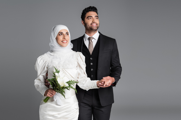 happy groom in suit holding hand of dreamy muslim bride with wedding bouquet of calla lily flowers isolated on grey - Foto, afbeelding
