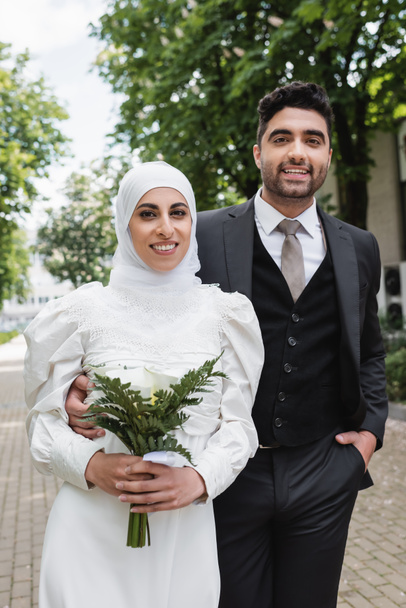 χαρούμενο γαμπρό ποζάρουν με το χέρι στην τσέπη κοντά muslim νύφη σε μαντίλα με γαμήλιο μπουκέτο  - Φωτογραφία, εικόνα