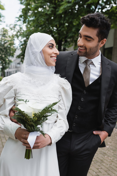 happy groom posing with hand in pocket and looking at muslim bride in hijab with wedding bouquet  - Foto, imagen
