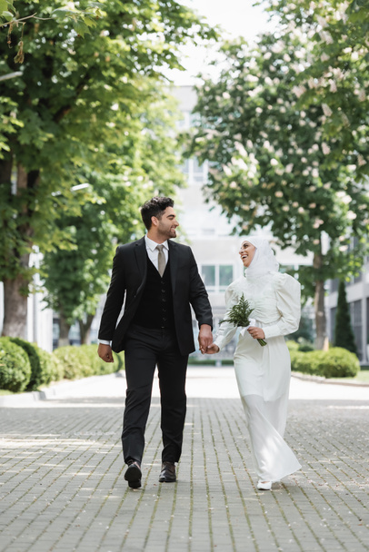 joyful groom holding hands with muslim bride with wedding bouquet and walking together  - Фото, изображение
