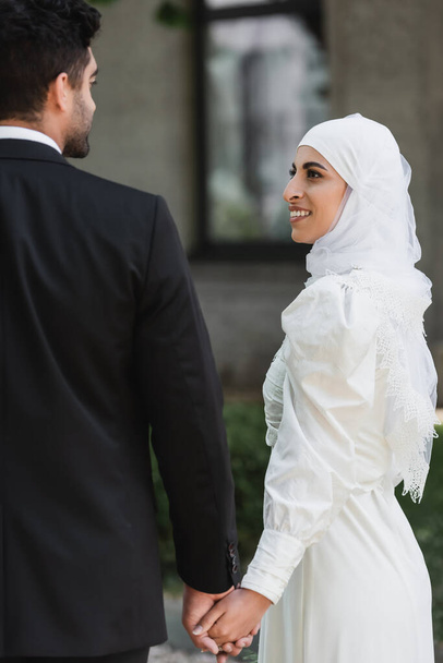 novio cogido de la mano con la novia musulmana sonriente en vestido de novia fuera   - Foto, Imagen