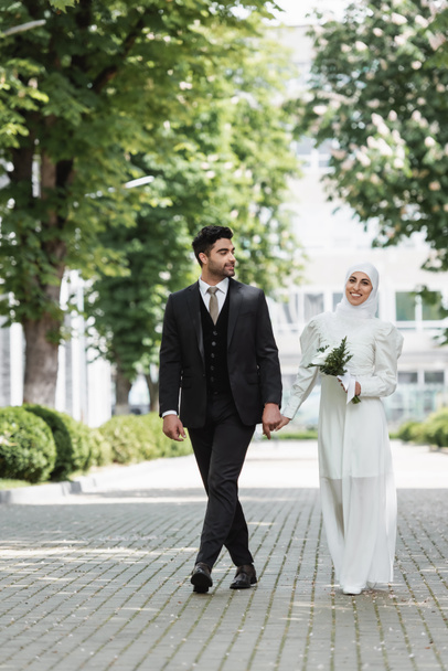 happy groom holding hands with smiling muslim bride in hijab with wedding bouquet  - Fotó, kép