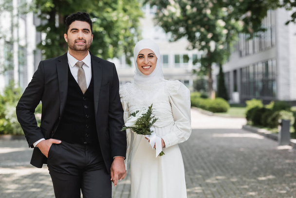 portrait of happy groom holding hands with smiling muslim bride in hijab with wedding bouquet  - Photo, Image