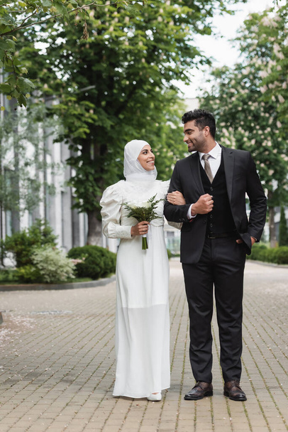 longitud completa de la novia musulmana feliz en hijab boda y vestido blanco que sostiene ramo cerca del novio fuera  - Foto, Imagen