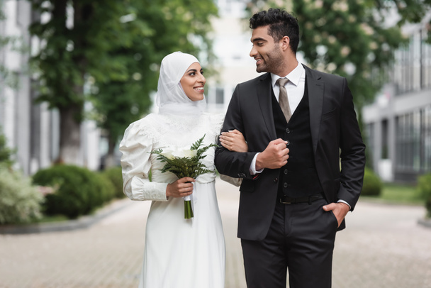 noiva muçulmano feliz no casamento hijab e vestido branco segurando buquê perto do noivo fora  - Foto, Imagem