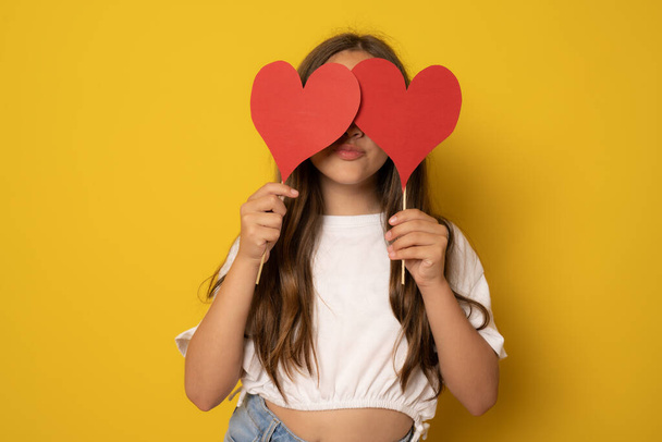 Girl covering eyes with two carton paper small little heart figures standing isolated over yellow background. Love concept. - Φωτογραφία, εικόνα