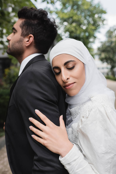 happy muslim bride in hijab and wedding dress hugging groom  - Foto, afbeelding