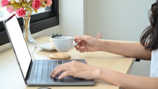Imagen recortada de la mano de la mujer sosteniendo la taza de café caliente y el uso de ordenador portátil. - Foto, Imagen