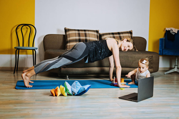 Fitness Side Hustles to Make Extra Cash. Personal Trainer Side Hustle. Young mother fitness instructor personal trainer doing online personal training near laptop at home with baby daughter - Photo, Image