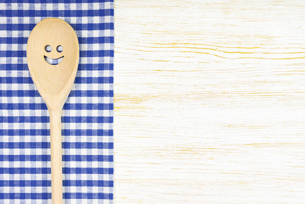 Wooden spoon with smile on blue checkered tablecloth on white wooden surface. Mockup for menu or recipe, restaurant, website with cooking. Kitchen food background, template, flat lay with copy space - Foto, afbeelding