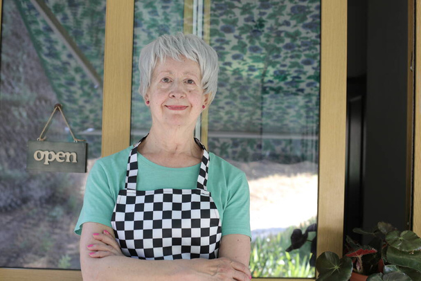 close-up portrait of mature woman in checkered apron on cafe door background - Foto, afbeelding