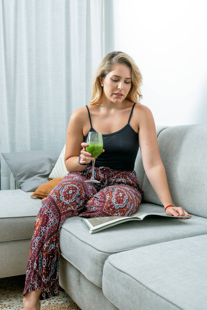νεαρή ξανθιά γυναίκα κάθεται στον καναπέ πίνοντας ένα χυμό λαχανικών - Φωτογραφία, εικόνα