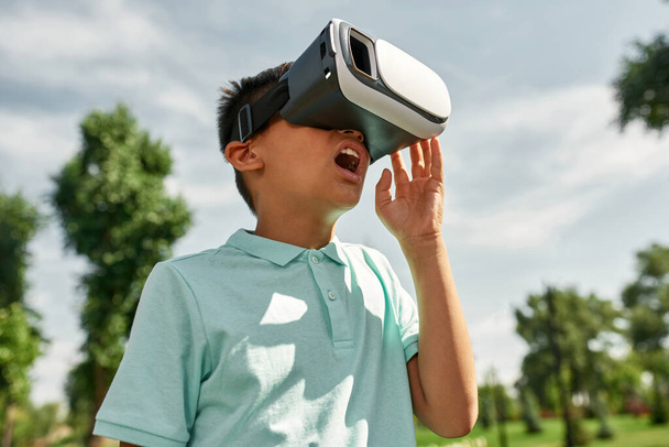 Excited shocked boy watching Virtual reality headset outdoors. Cropped dark-haired male child of digital generation alpha. Modern childhood lifestyle. Gadget addiction. Green park in sunny summer day - Foto, Imagen