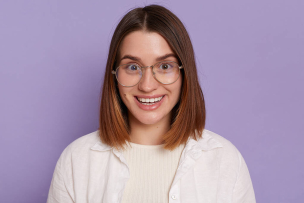 Indoor shot of attractive young woman wearing white clothing and glasses posing isolated over purple background, looking at camera with happy positive expression. - Foto, afbeelding