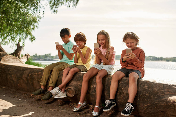 Group of multiracial children sitting on log and using smartphones on river coast outdoors. Boys and girl of generation alpha. Gadget addiction. Childhood lifestyle. Sunny summer day - Φωτογραφία, εικόνα