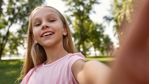 Cropped image of smiling caucasian girl looking at camera during taking selfie in blurred park. Beautiful blonde female child of generation alpha. Concept of childhood lifestyle. Sunny summer day - Photo, Image