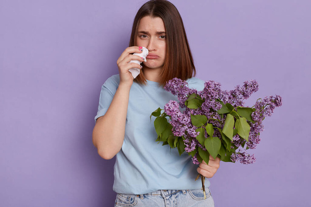 Indoor shot of Caucasian unhealthy woman wearing blue t shirt suffering from runny nose, having allergic rhinitis reaction on lilac, posing isolated over purple background. - Foto, imagen