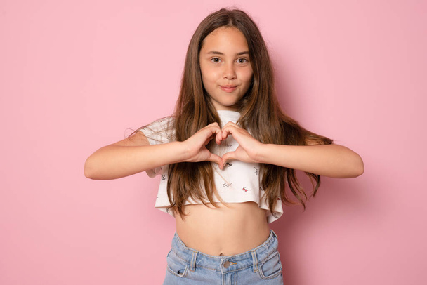 Pembe arka planda izole edilmiş tişört giyerek elleriyle kalp formu oluşturan güzel bir genç kızın portresi. - Fotoğraf, Görsel