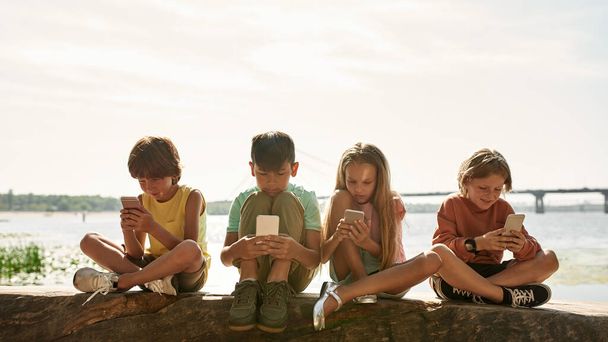 Group of multiethnic children sitting on log and browsing smartphones on river coast outdoors. Boys and girl of generation alpha. Gadget addiction. Childhood lifestyle. Sunny summer day - Foto, Imagem