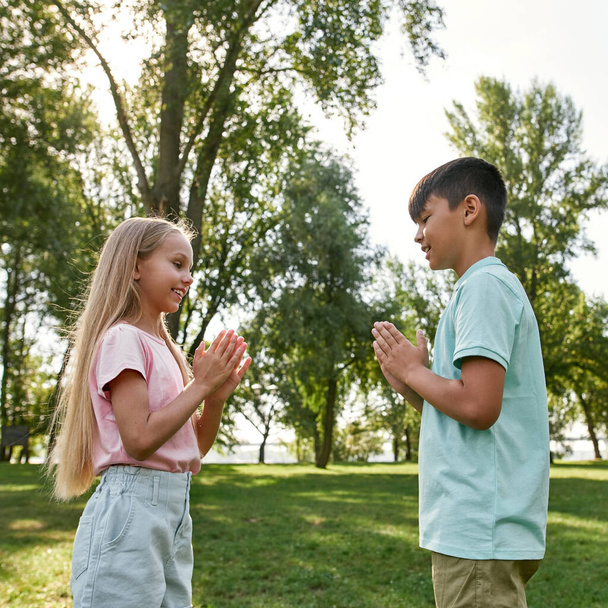 Side view of smiling caucasian girl and multiracial boy playing patty cake game on green meadow in blurred park. Beautiful children of generation alpha. Childhood lifestyle. Friendship. Sunny day - Foto, Imagen