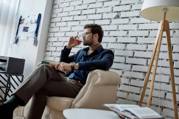 Successful caucasian IT investor or seller with book drinking cognac or brandy and thinking in armchair in office. Business strategy, development, promotion, selling and marketing of high-tech company - 写真・画像